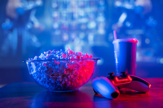 Video Games On Big Screen TV. Popcorn In A Large Bowl, A Soda Drink In A Plastic Cup And A Joystick. Neon Lighting. Vacation With Friends, Online Competitions, Games, Fast Food.