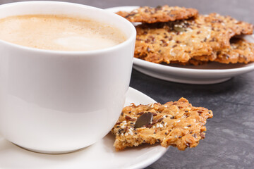 Cup of coffee with milk and fresh baked oatmeal cookies with honey and healthy seeds on white plate. Delicious crunchy dessert