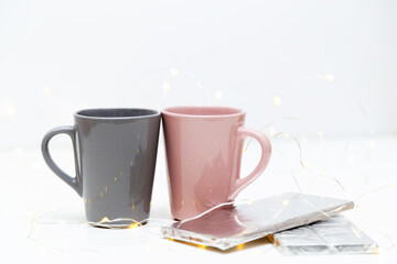 Two pink and grey coffee mugs mockup and a bar of chocolate on white background with small golden lights and bokeh. Blank template for your design, branding, business