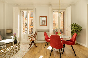 Living room in apartment with metal and glass side table, two-seater sofa upholstered in gray fabric, round marble dining table, chairs upholstered in red velvet fabric and balconies with white doors
