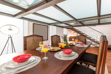 Studio living room with rectangular wooden dining table with lots of assorted fruits, glasses of juice and metal and translucent glass ceiling