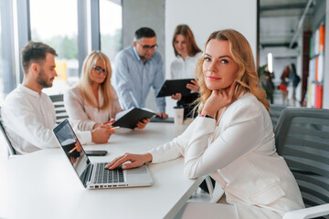 Mature woman is sitting by the table with laptop. Group of professional business people is in the office