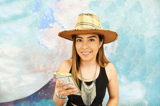 Mujer joven latina con sombrero, sonriente y usando su telefono movil.