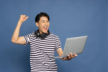 Happy young Asian pro gamer with headphones holding computer laptop and win in online video game...