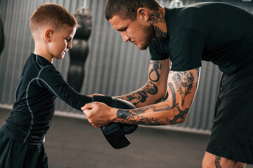 Man is helping child to wear the glowes. Coach is teaching the boy box techniques indoors