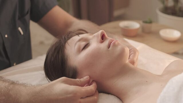 Slowmo closeup of unrecognizable masseur massaging ear lobes of beautiful young woman lying with eyes closed on couch in spa salon
