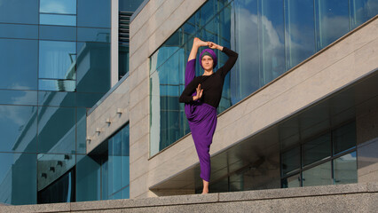 Strong muslim woman in hijab flexible young girl stretching yogi yoga workout tree pose asana female raises leg in air doing split stretch one foot balance standing city urban building background