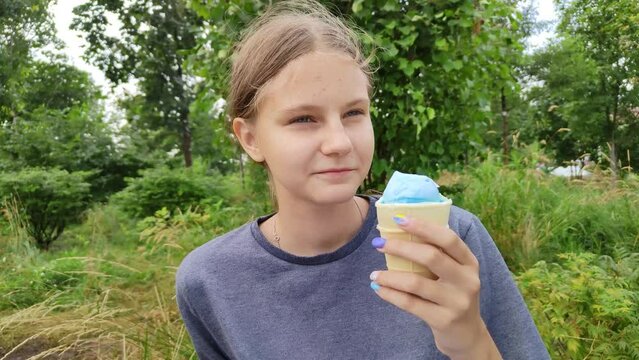 Teen girl eats turquoise ice cream. Young beautiful woman with colorful nails walks in city park on summer day. Child eats sweets. Cold ice cream on hot day. Teenager in a garden, park or forest