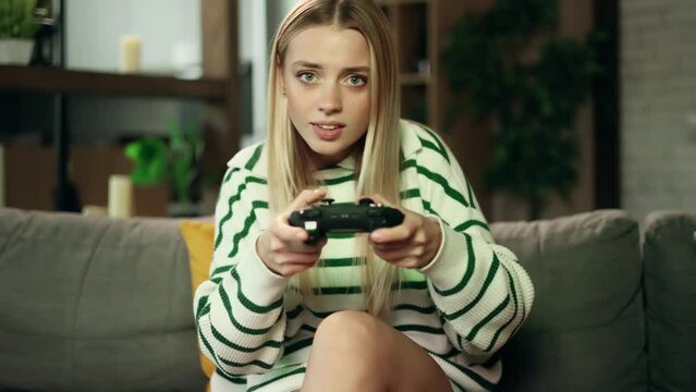 Beautiful excited young gamer girl sitting on a couch and playing in video games on a console. Happy player woman controlling joystick for videogame.