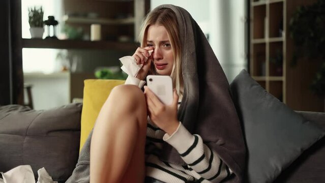 Crying young woman with phone sitting on the couch and reading bad news email, break up message from ex-boyfriend. 