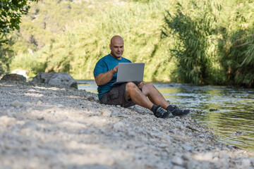 Digital nomad man working with a laptop and a phone, sitting on the bank of a river. 