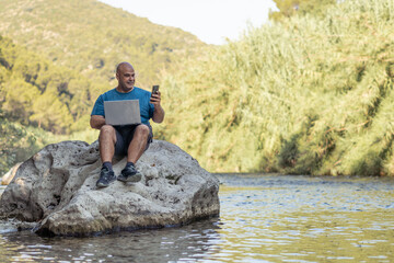 Digital nomad man working with a laptop sitting on a rock above the water of a beautiful and calm river.