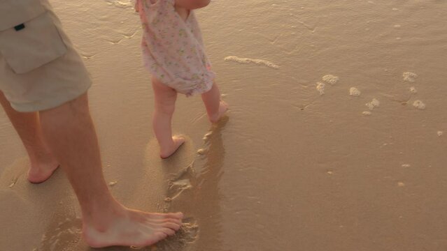 Close up male child barefoot legs stepping in wet water ocean sea play with waves. Unrecognizable father holding baby daughter toddler by hand helping support kid in first steps walking on sand beach
