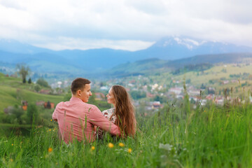 Romantic couple of young people sitting on the grass in field of spring flowers with mountain views in the background. Man and woman are looking at each other.