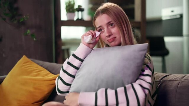 Young attractive woman sits on sofa at home alone, hugging a pillow and watch drama film. Beautiful girl wipes away tears after watching film.
