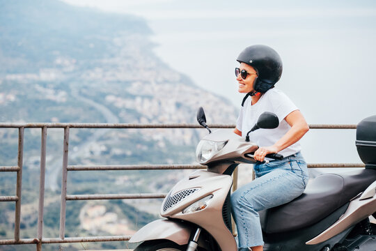 Middle-age woman in helmet and sunglasses on motor scooter on the Sicilian old town streets in the Forza d'Agro with Sant'Alessio Siculo harbor. Happy Italian vacation and transportation concept.