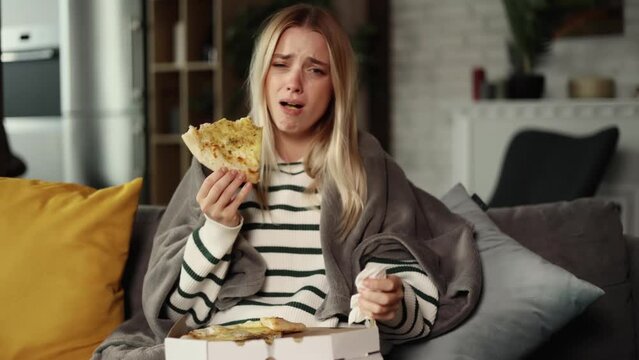 Sad young woman dealing with stress by eating food. Upset young woman wrapped in blanket, crying and eating pizza.
