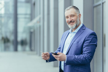 Obraz na płótnie Canvas Portrait of successful gray-haired businessman man smiling and looking at camera, boss holding bank credit card and smartphone, director using money transfer app