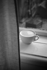 A cup of cappuccino on the windowsill behind a curtain. black and white