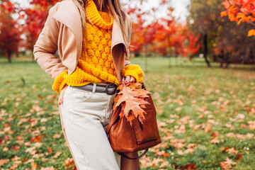 Close up of stylish woman holding purse wearing yellow sweater in autumn park. Fall female clothes, accessories. Fashion
