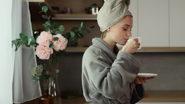 Young beautiful woman in towel on head looks out the window, drinks a hot drink of tea or coffee. Beautiful morning.