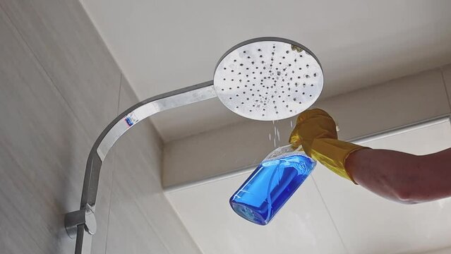 The hand of a caucasian young woman in yellow gloves holds out a transparent bottle with blue liquid and sprays on a large round shower head in the bathroom,close-up view from below.The concept of cle