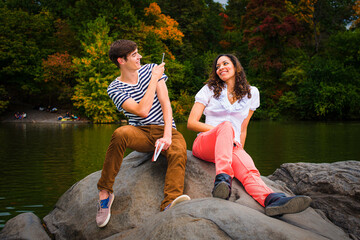 Young couple making fun by a lake in a park. Guy wearing a striped t shirt, brown pants, sneakers, holding a book, taking a picture. Girl dressing in white top, red pants, boot shoes, smiling. .