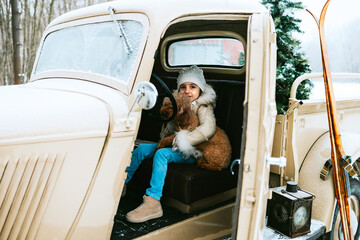 cute beautiful little Armenian girl in winter coat and knitted hat and pet poodle dog sit near beige retro pickup truck decorated for Christmas and New Year vintage interior suitcases, skis, sledge © klavdiyav