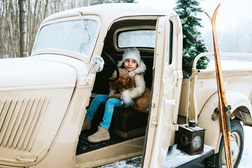 cute beautiful little Armenian girl in winter coat and knitted hat and pet poodle dog sit near beige retro pickup truck decorated for Christmas and New Year vintage interior suitcases, skis, sledge