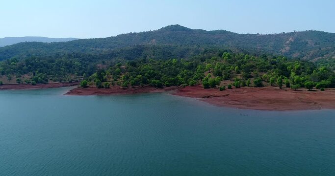 Aerial shot of river running along forest with reddish soil, hill background