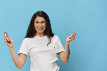 portrait of a beautiful, pleasant woman with long hair, in a light T-shirt, holding a fashionable smartphone in her hands and joyfully raising her hands up. Horizontal photo on a light blue background