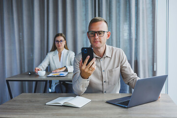 Fototapeta premium A male manager is talking on a mobile phone while sitting at a table with a computer and a notepad. Working atmosphere in an office with large windows