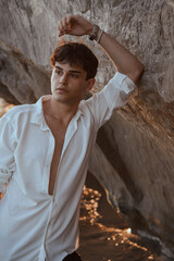 Portrait of handsome sexy young man in white shirt posing staying near sand stones on the beach near sea at sunset.