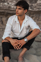 Portrait of handsome sexy young man in white shirt sitting on the sand stones on the beach near sea...