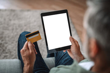 Retired european man with beard uses credit card and tablet with empty screen, shopping online in living room