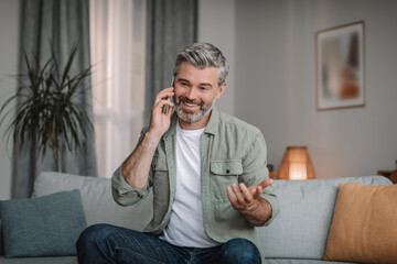 Smiling handsome retired european man with beard calls by smartphone, talk remote in living room...
