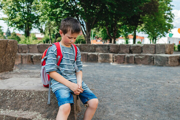 A sad schoolboy sits in the school yard with a backpack on his back. Children's problems at school. The boy does not want to go to school