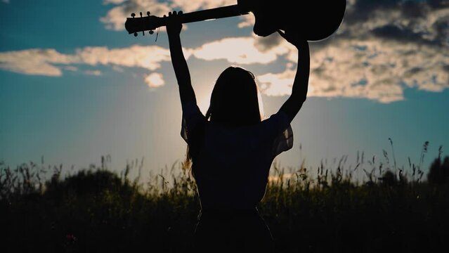 Silhouette of a girl with a guitar that she holds over her head. She walks across the field towards sunset
