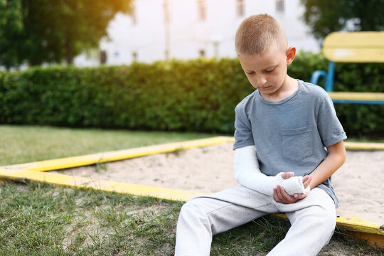 child caucasian with broken limb outdoors sits near the playground and looking on the plaster bandage on his arm. the worst summer vacation. concept of health accident, and medical