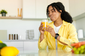 Young african american woman drinking fresh orange or mango juice from glass, sitting in kitchen,...