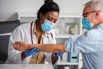 Female doctor is examining male elderly patient at clinic. Doctor and patient wearing a face mask.