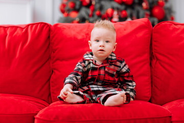 Naklejka premium adorable little baby boy in a red christmas outfit sitting on red sofa in front of a christmas tree. Beautiful little baby boy celebrating Christmas.Family on Christmas eve at fireplace.
