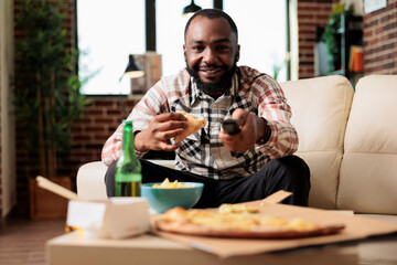 African american man using tv remote contorl to switch channel and eating slice of pizza from fast food takeout delivery. Having fun with takeaway meal and beer while he watches movie.