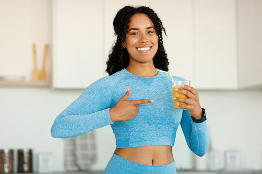 Happy african american lady in sportswear pointing finger at glass of orange juice, standing in kitchen and smiling