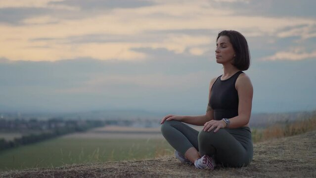 Young beautiful woman looking at the smartwatch while sitting at the lotus yoga position on a grassy hill during the evening