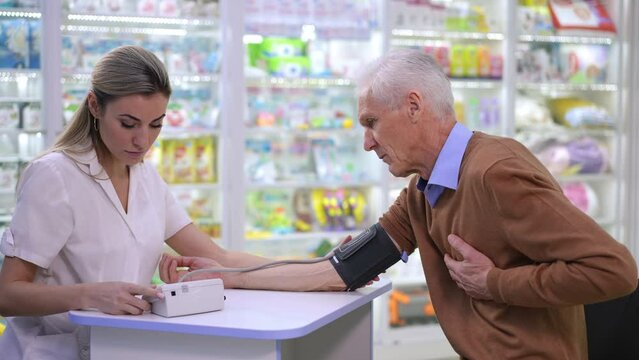 Side view pharmacist measuring blood pressure of unwell senior client in drugstore. Concentrated intelligent young woman helping ill Caucasian man holding chest sitting in pharmacy