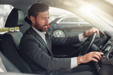 Handsome businessman in grey suit is riding behind steering wheel of car and turns on music