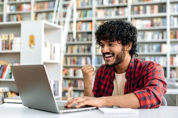 Cheerful amazed indian or arabian guy, student, sitting in university library with laptop, rejoices in success, win, good mark on the exam, gesturing hand, smiling, emotional face expression