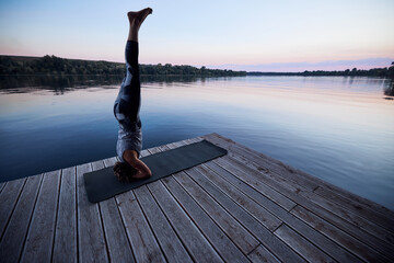 A fit woman practices yoga on the dock near the river and balances on her head in the Supported...