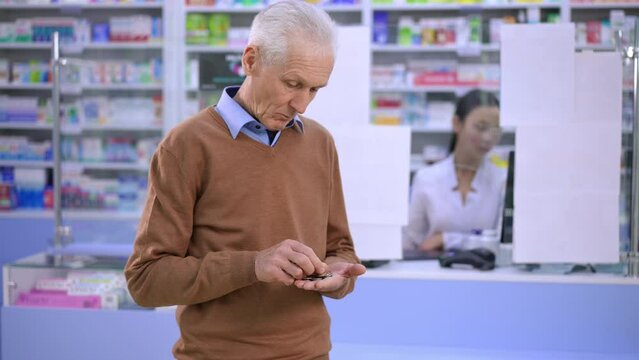 Sad poor senior Caucasian man counting coins standing in pharmacy looking back at cash desk with blurred Asian cashier. Portrait of moneyless frustrated client in expensive drugstore indoors